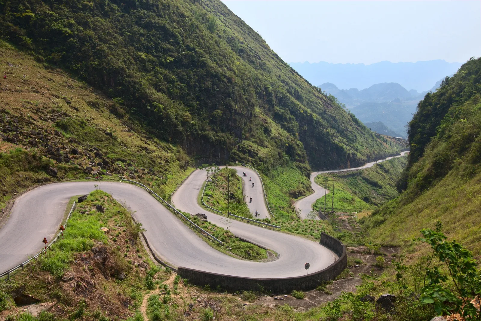 A winding mountain road switchbacks through green hillsides in Ha Giang province.