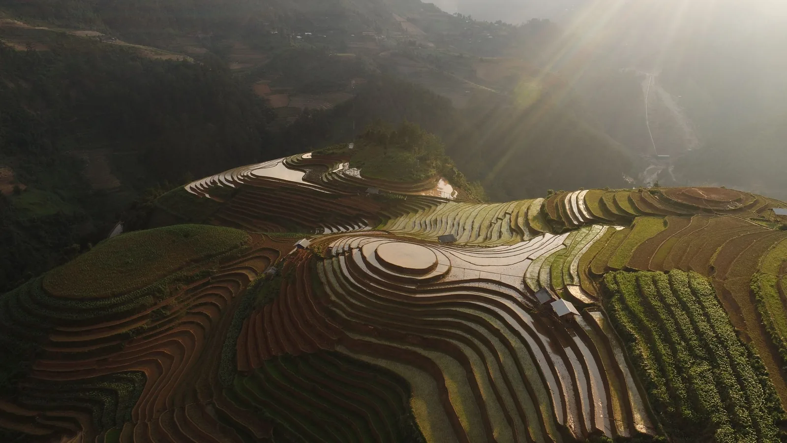 Sunlight streaks over water-filled terraced rice fields carved into the hillsides of Mu Cang Chai.