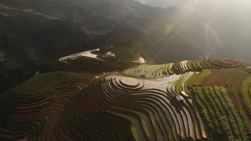 Sunlight streaks over water-filled terraced rice fields carved into the hillsides of Mu Cang Chai.
