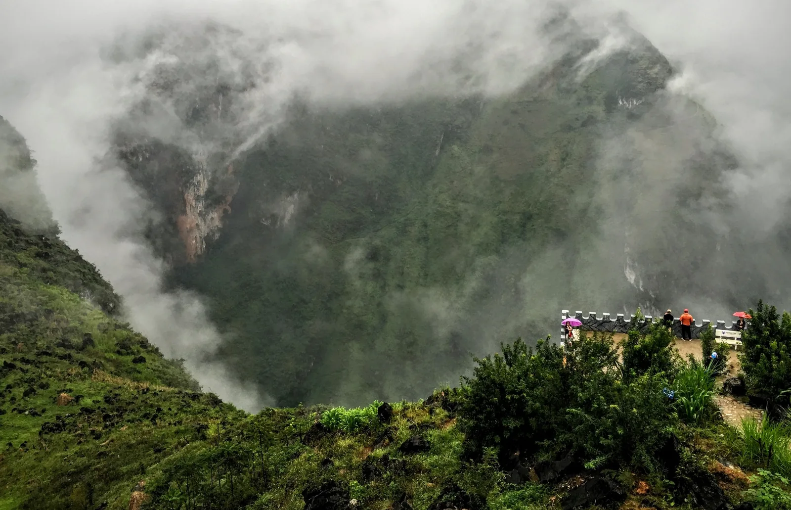 Mist swirls around a deep green valley at Ma Pi Leng Pass in Ha Giang, with a small viewing platform on the ridge.