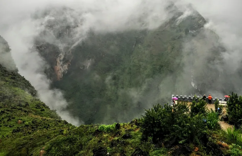 Mist swirls around a deep green valley at Ma Pi Leng Pass in Ha Giang, with a small viewing platform on the ridge.