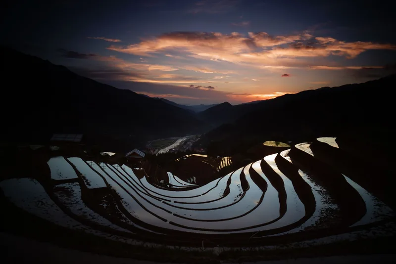 Water-filled rice terraces curve down a mountainside at dusk in Mu Cang Chai under a glowing sunset sky.