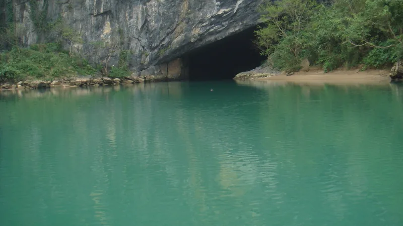 A dark cave mouth opens onto a turquoise river beneath limestone cliffs at Phong Nha.