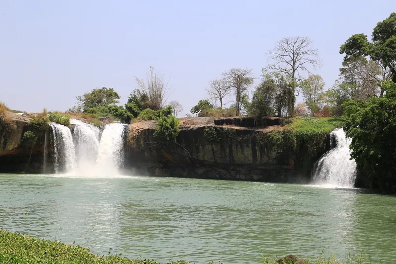 Multiple cascades of Dray Nur waterfall plunge over a wide rock ledge into a calm green river in Dak Lak.